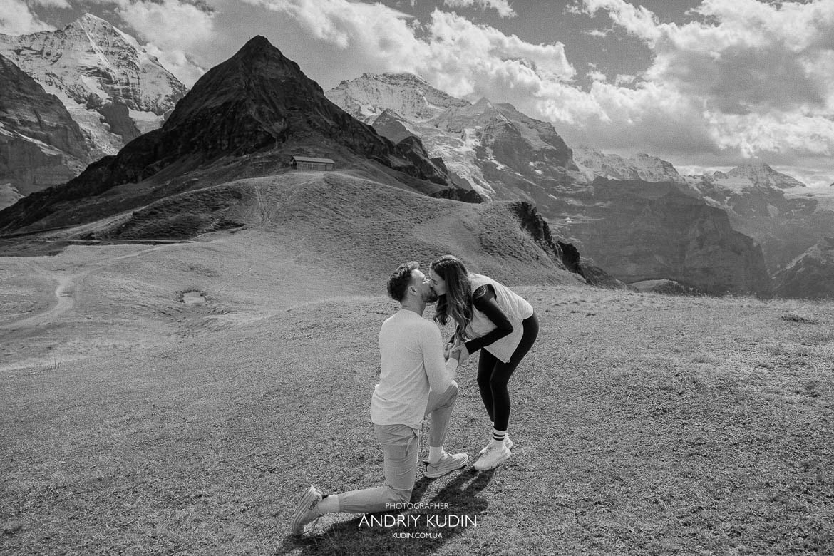 Man kneeling with engagement ring on a quiet meadow above Grindelwald with Eiger peak in sight.