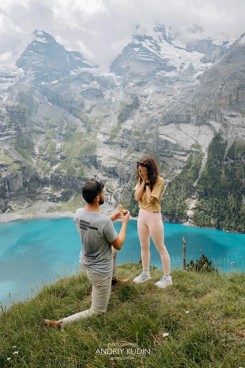 Man kneeling during an Oeschinensee proposal on a grassy cliff above the turquoise lake, captured by a Switzerland proposal photographer. Man kneeling during an Oeschinensee proposal on a grassy cliff above the turquoise lake, captured by a Switzerland proposal photographer.