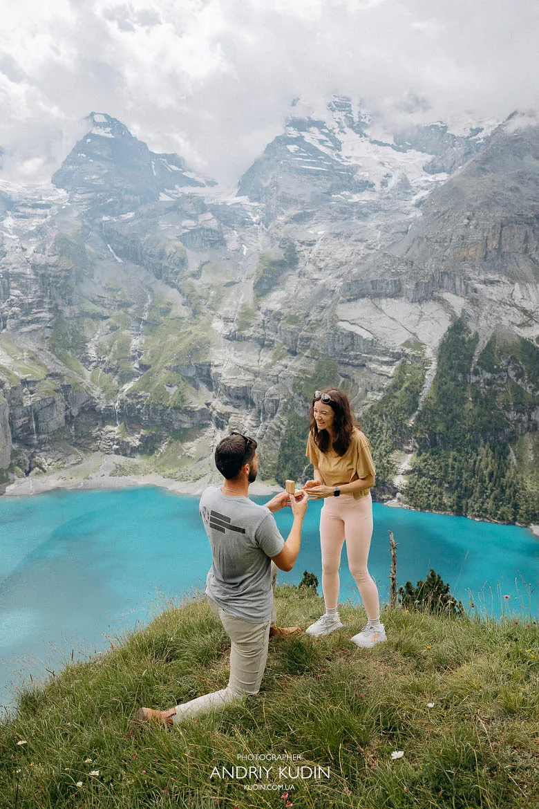 Man kneeling during an Oeschinensee proposal on a grassy cliff, captured by a Switzerland proposal photographer at Lake Oeschinen. Man kneeling during an Oeschinensee proposal on a grassy cliff, captured by a Switzerland proposal photographer at Lake Oeschinen.