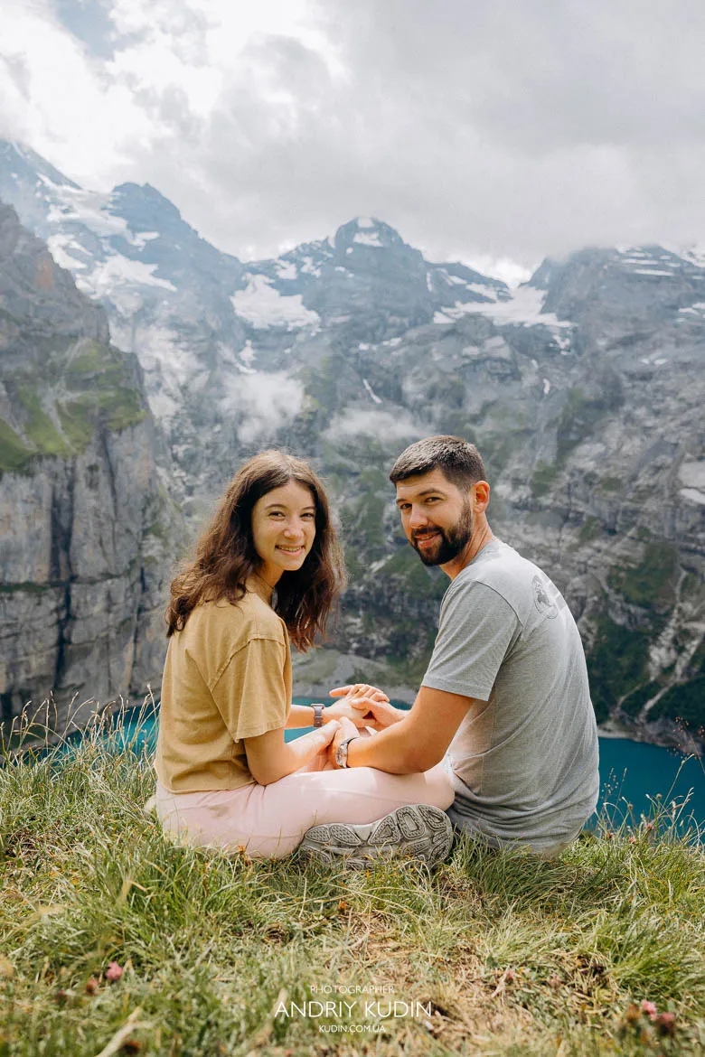 Lake Oeschinen proposal moment with a couple sitting close together on a grassy viewpoint overlooking the lake and rocky peaks. Lake Oeschinen proposal moment with a couple sitting close together on a grassy viewpoint overlooking the lake and rocky peaks.