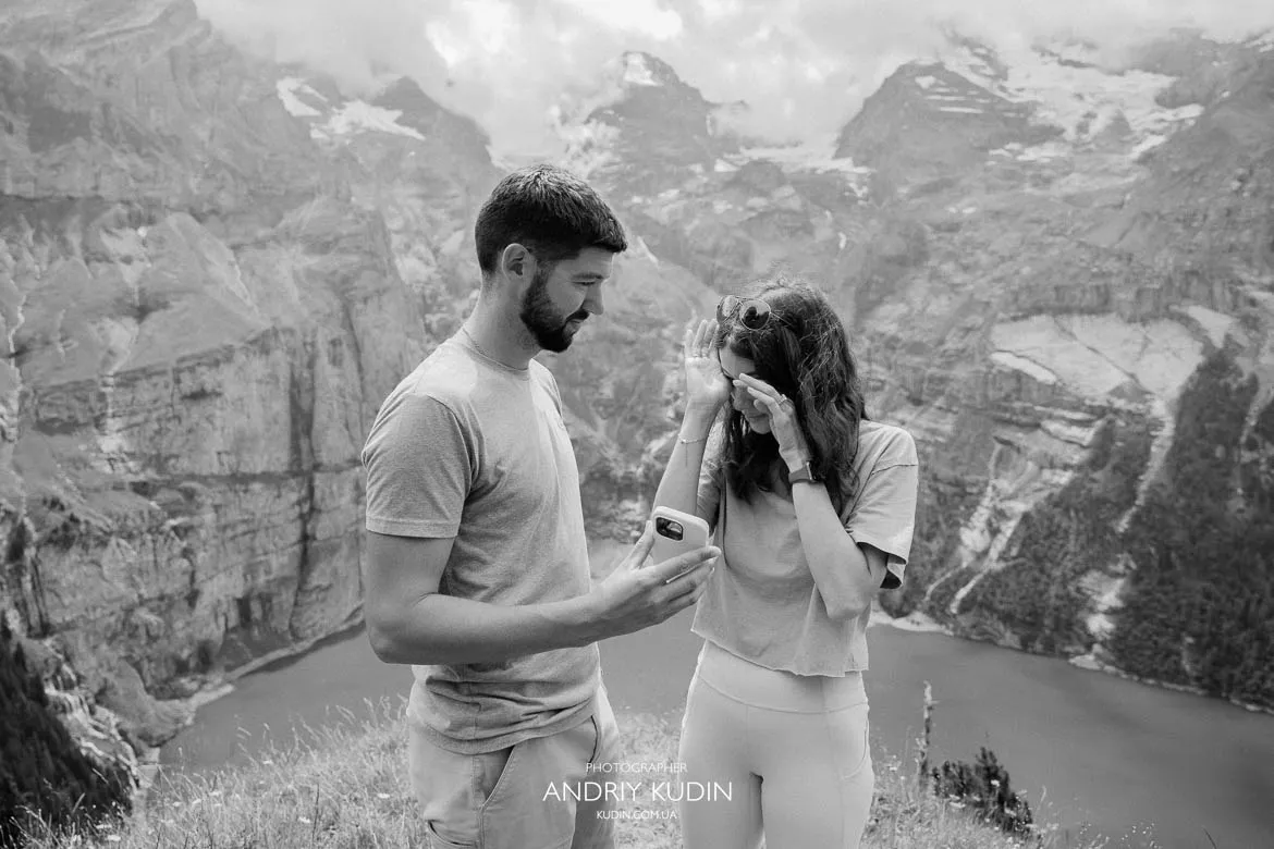 Marriage proposal photography at Lake Oeschinen showing a couple checking a phone on a grassy viewpoint with alpine cliffs in Switzerland.