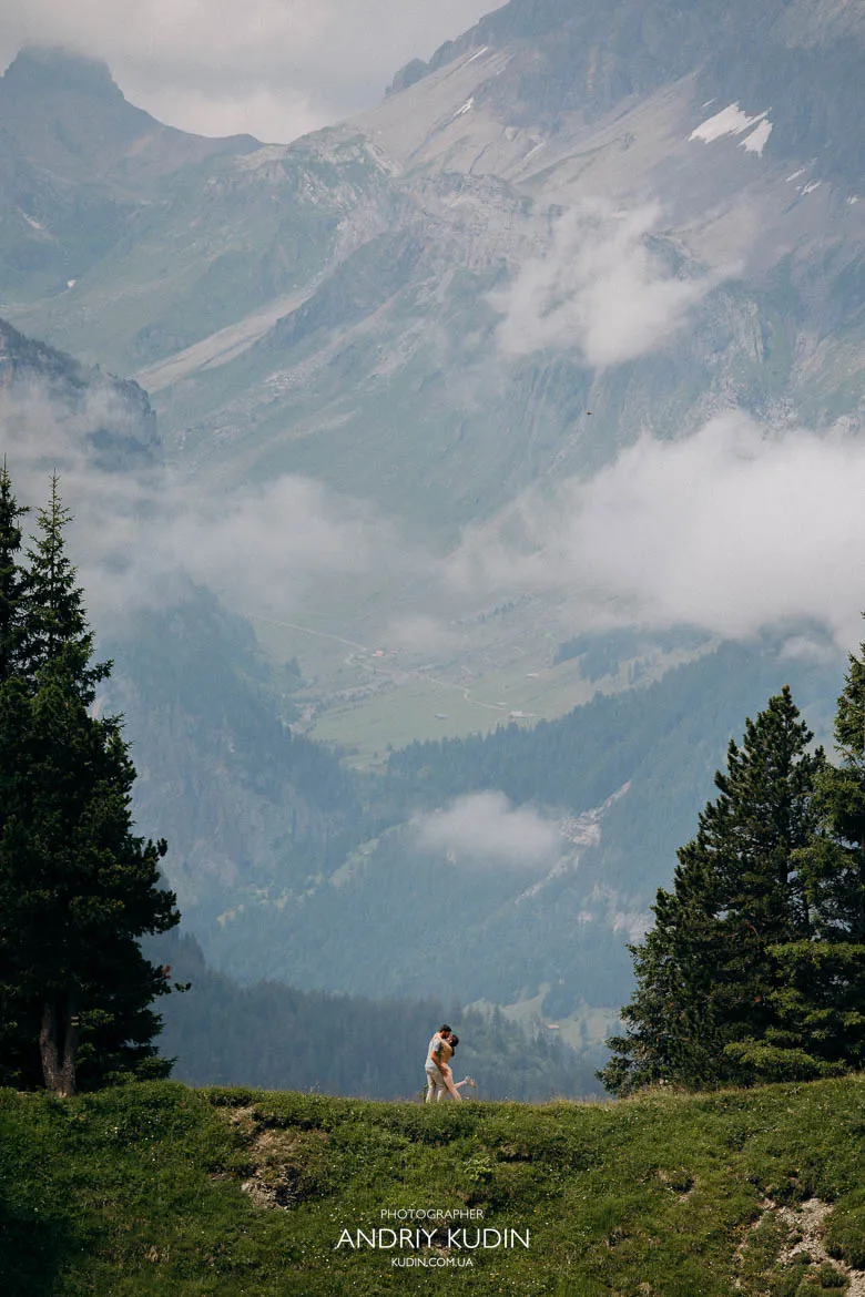 Couple standing on a grassy ridge above Lake Oeschinen, surrounded by trees and misty alpine peaks Couple standing on a grassy ridge above Lake Oeschinen, surrounded by trees and misty alpine peaks