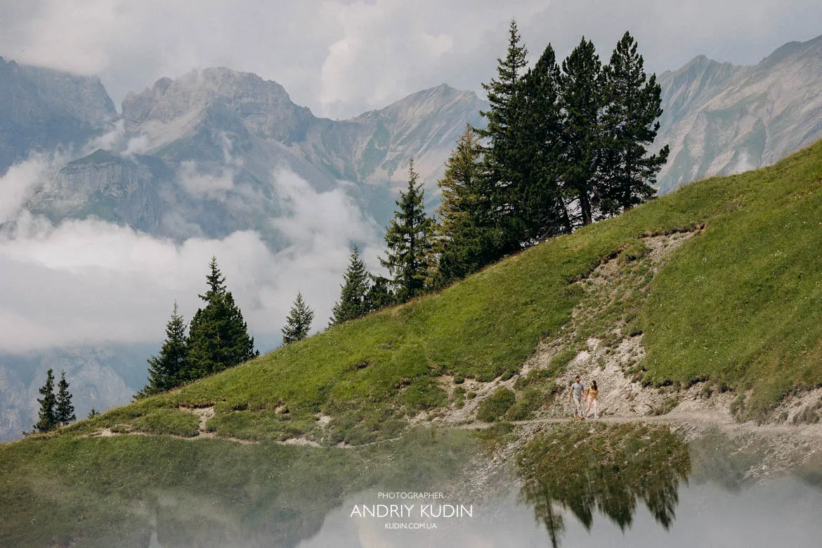 Couple walking along a grassy ridge above Lake Oeschinen with evergreen trees, misty peaks and reflections in a calm alpine pond. Couple walking along a grassy ridge above Lake Oeschinen with evergreen trees, misty peaks and reflections in a calm alpine pond.