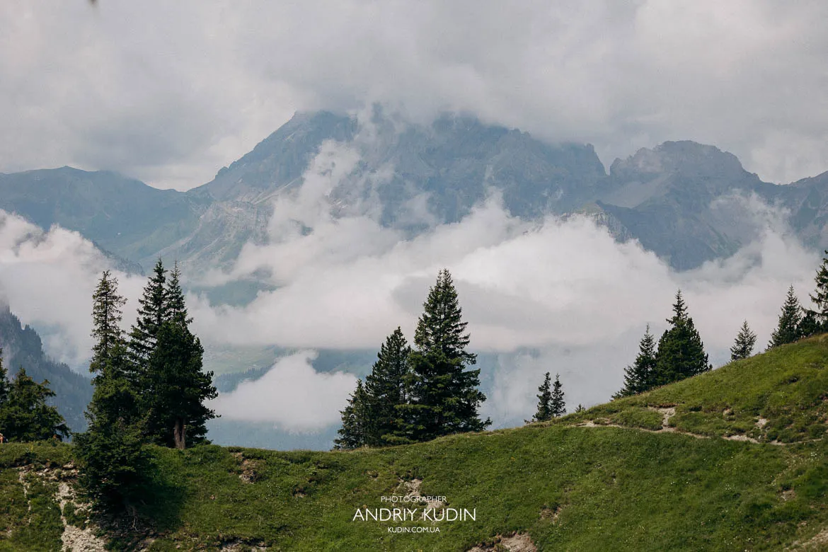 Alpine scenery near Lake Oeschinen showing grassy slopes, scattered trees and cloud‑covered mountain peaks used for marriage proposal photography.
