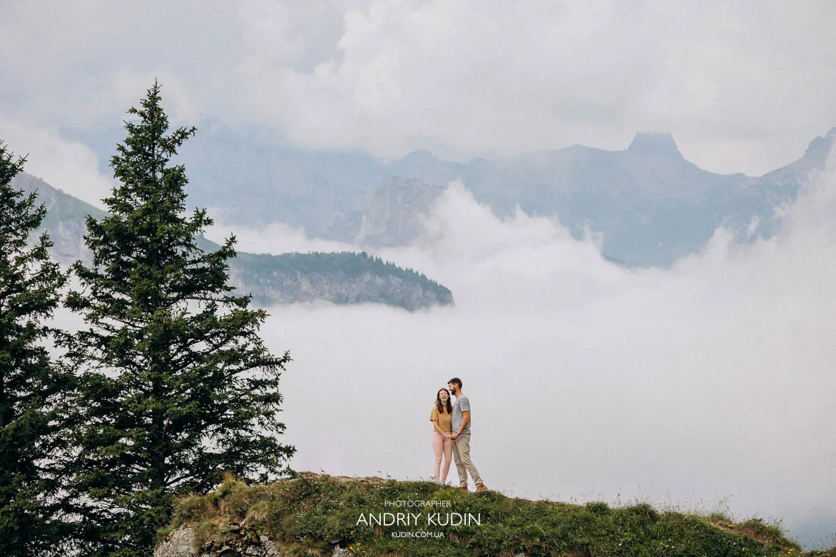 Couple standing close together on a misty hilltop above Lake Oeschinen with evergreen trees and fog‑covered alpine peaks in Switzerland. Couple standing close together on a misty hilltop above Lake Oeschinen with evergreen trees and fog‑covered alpine peaks in Switzerland.