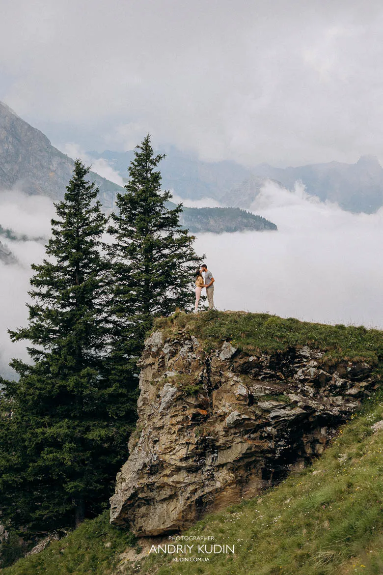 Couple standing on a rocky cliff above Lake Oeschinen, surrounded by evergreen trees and misty alpine peaks in Switzerland. Couple standing on a rocky cliff above Lake Oeschinen, surrounded by evergreen trees and misty alpine peaks in Switzerland.