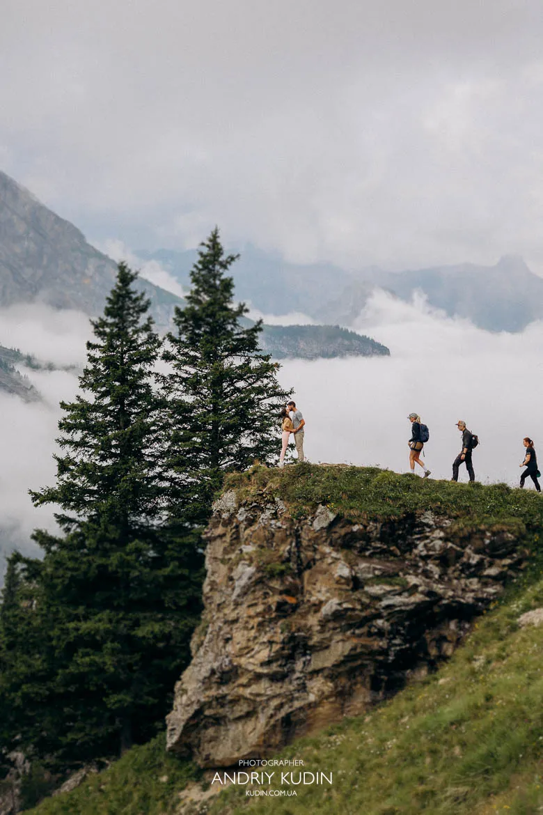 Hikers exploring a grassy cliffside above Lake Oeschinen, surrounded by alpine trees, rocky slopes and misty mountain peaks. Hikers exploring a grassy cliffside above Lake Oeschinen, surrounded by alpine trees, rocky slopes and misty mountain peaks.