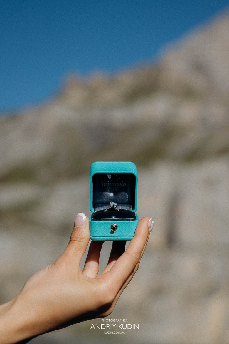Man proposing with a Tiffany engagement ring beside Lake Oeschinen’s turquoise water and mountain backdrop.