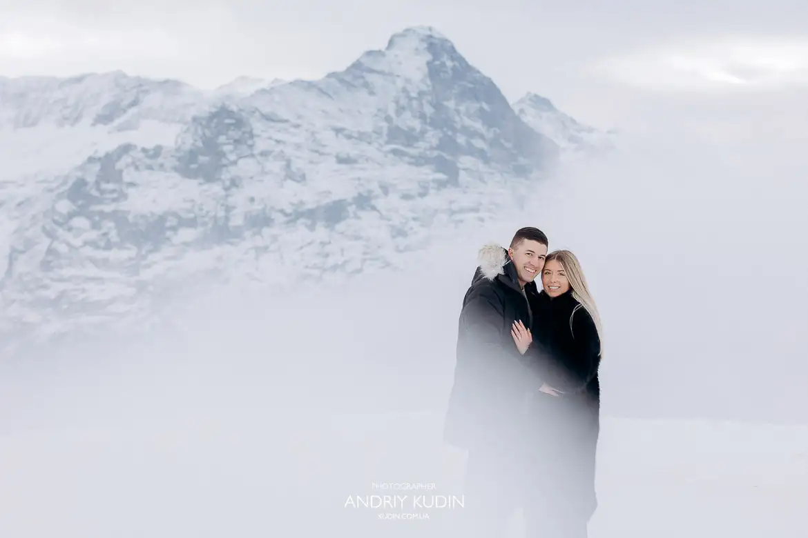 The couple in warm jackets celebrates their engagement on the thrilling Cliff Walk suspension bridge at Grindelwald First, Switzerland, with a deep gorge and mountains below.