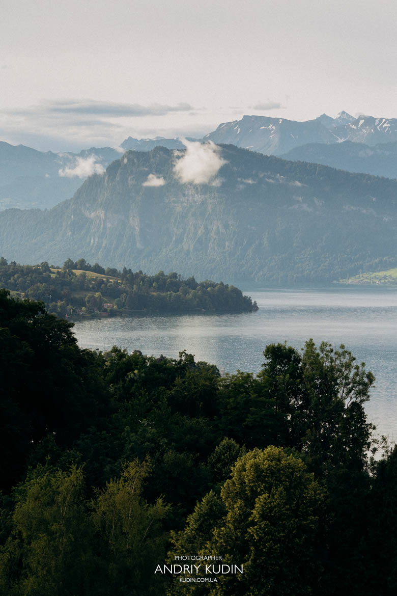 Scenic overlook of Lucerne old town roofs and towers during a sunrise proposal shoot. Cinematic mountain range behind a couple standing in an elevated park above Lucerne.