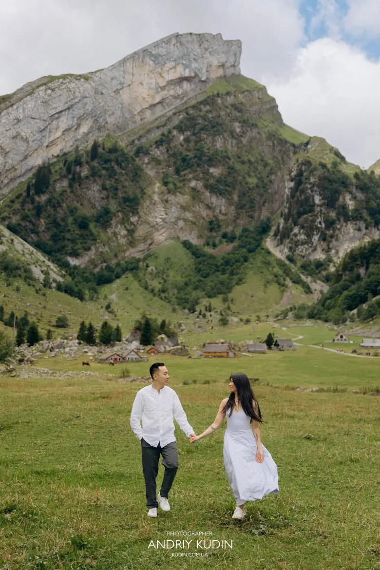Engaged couple walking hand in hand across green alpine meadow at Seealpsee summer photoshoot