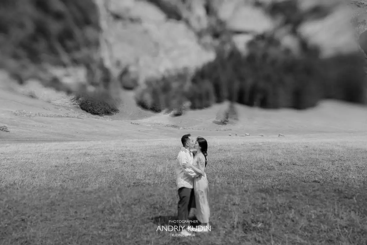 Black and white wide shot of couple kissing in open alpine meadow in Swiss Alps captured by proposal photographer