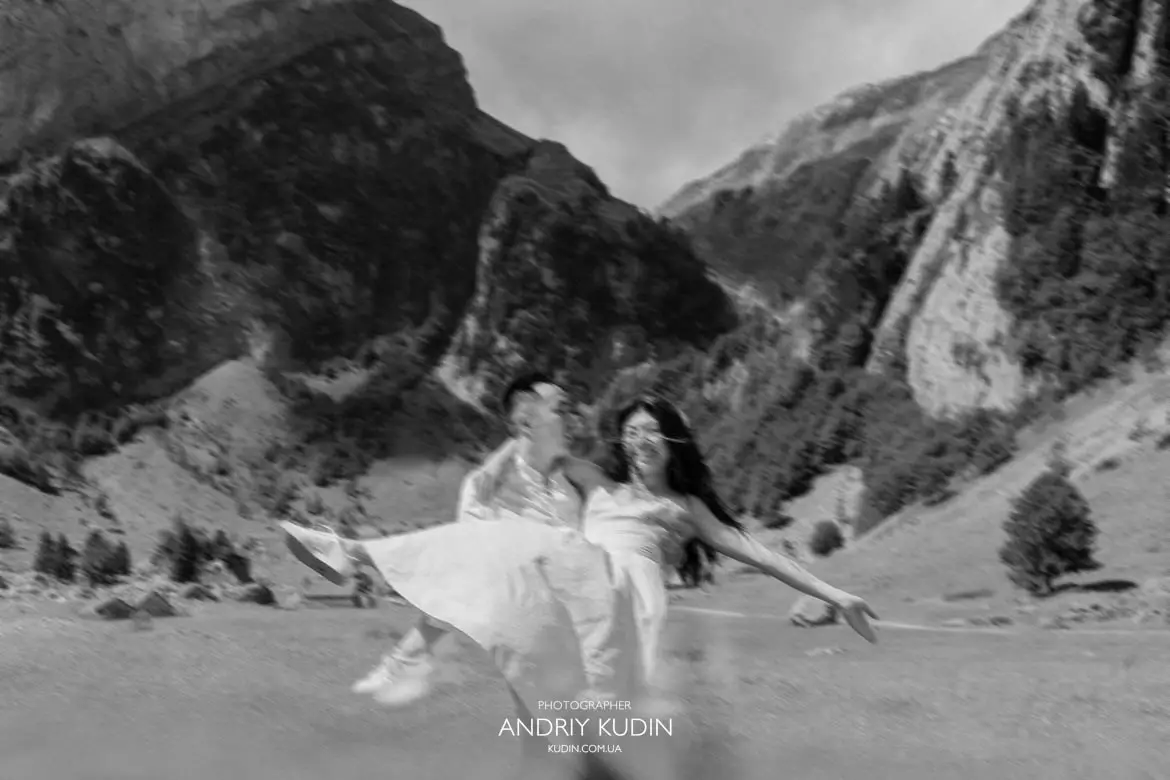 Joyful black and white action shot of man carrying fiancé in his arms on alpine field after engagement