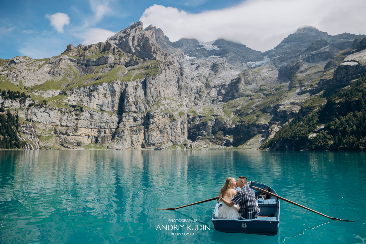 Wide panoramic view of couple kissing in a rowboat on turquoise waters of Lake Oeschinen surrounded by mountain cliffs