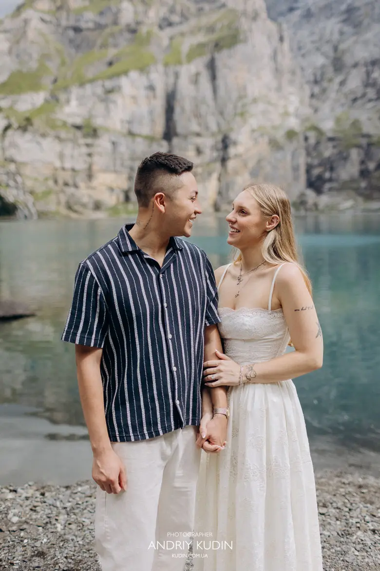 Smiling engaged couple holding hands by Lake Oeschinen shore after successful marriage proposal in Swiss Alps
