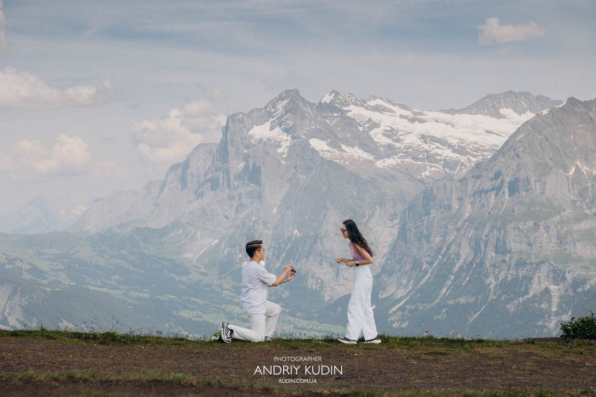 Man kneeling on one knee proposing to his girlfriend on Mannlichen summit with Swiss Alps background